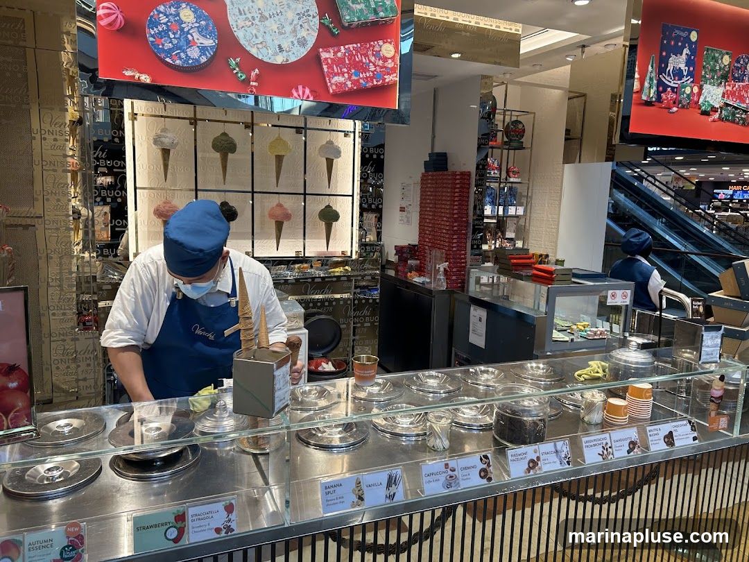 A person in a blue apron and hat is preparing ice cream behind a counter. The counter displays various flavors of ice cream in metal containers. There are also labels with flavor names and descriptions. In the background, there are displays of packaged chocolates and ice cream cones. A shopping mall escalator is visible in the far background.