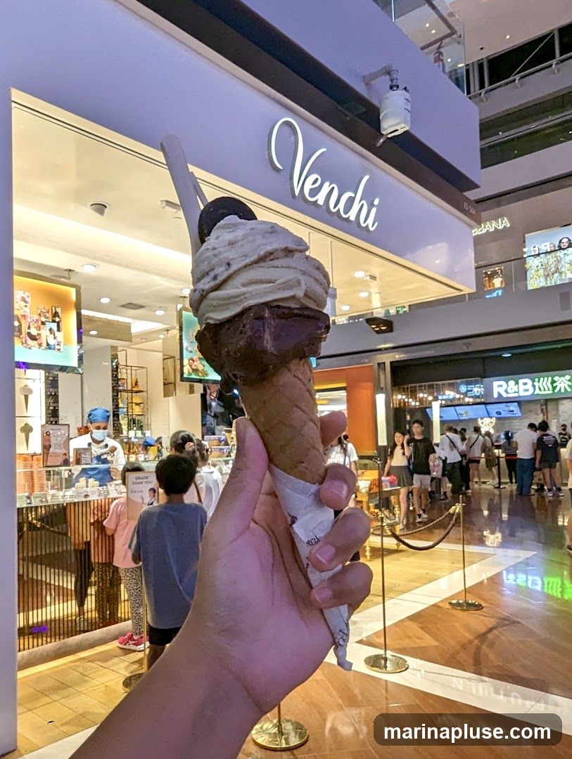 A hand holds a large ice cream cone with two scoops, topped with an Oreo cookie, in front of a Venchi store. The store has a prominent white sign with the name "Venchi" in cursive script. Inside the store, staff members are visible behind a counter, serving customers. The background shows a busy shopping mall with other shops and people walking around.