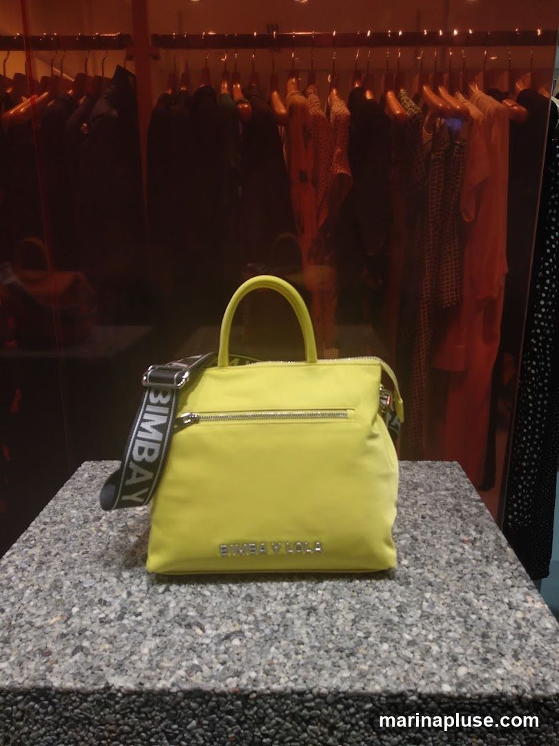 A bright yellow handbag with a black strap is displayed on a speckled counter. The strap has the word "BIMBA Y LOLA" printed on it in white. The handbag also has "BIMBA Y LOLA" embossed on its front. Behind the handbag, clothing items are visible on hangers in a dimly lit display area.