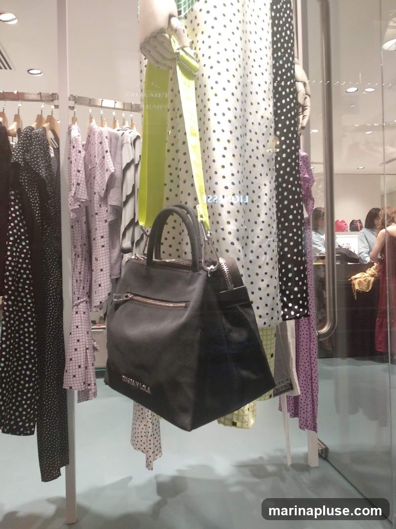 A store window display features a black handbag with a lime green strap hanging in front of polka dot and patterned dresses. The handbag has the brand name "BIMBA Y LOLA" embossed on it. In the background, through the glass, people can be seen inside the store.