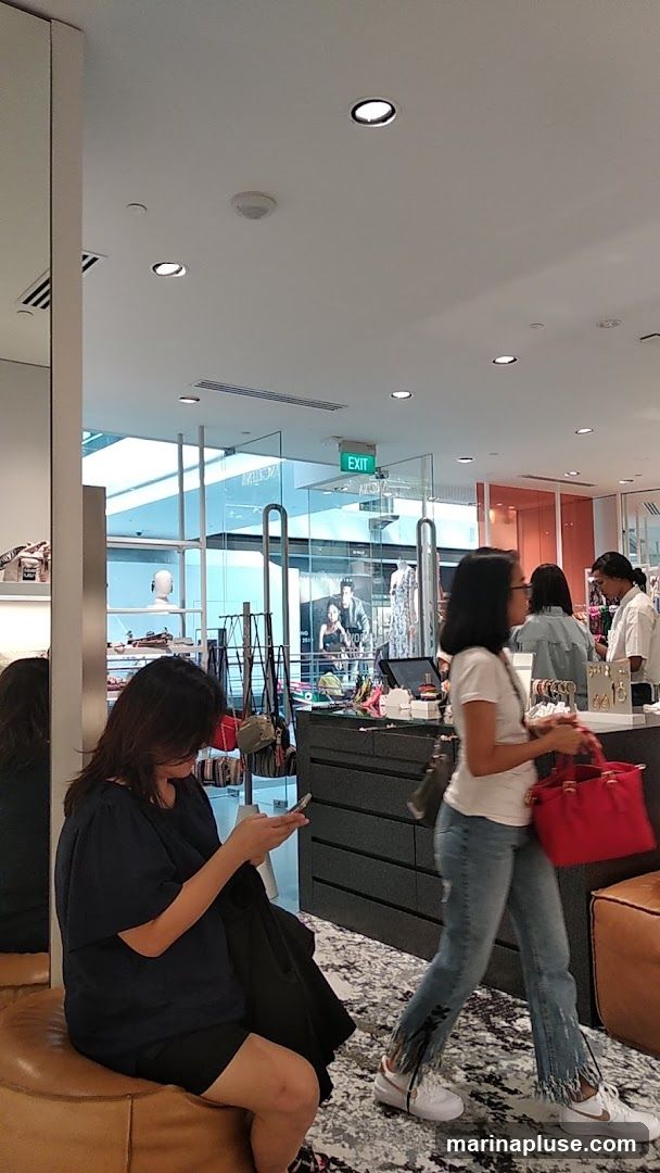 An interior view of a department store with several shoppers. In the foreground, a woman sits on a leather ottoman, looking at her phone. To her right, another woman walks by, carrying a red handbag. In the background, displays of clothing and accessories are visible, along with an "EXIT" sign.