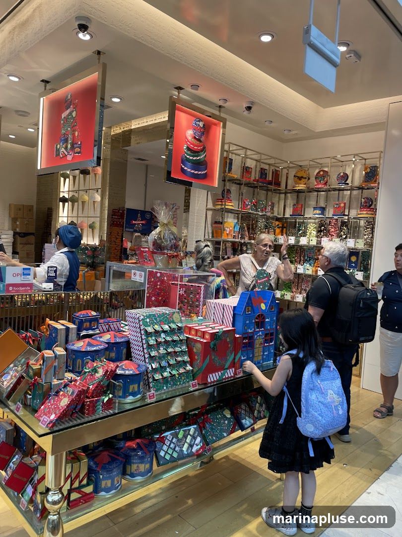 A store interior filled with festive Christmas-themed gift boxes and displays. A young girl with a backpack is looking at a display of advent calendars. Several adults are browsing the merchandise. The store appears to be a confectionery or gift shop, with colorful packaging and decorative items.