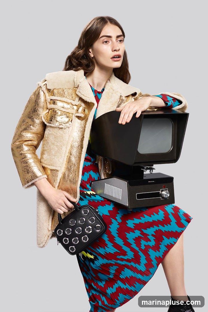A fashion model poses with a vintage camera and a black handbag. She is wearing a gold shearling-lined jacket over a patterned dress. The background is a plain gray.
