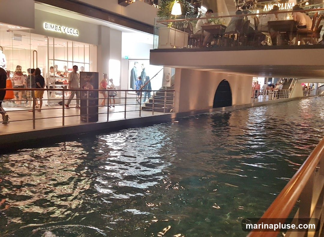 An indoor canal runs through a shopping mall, with shops and restaurants visible on either side. The BIMBA Y LOLA store is prominently displayed on the left, featuring mannequins in the window. On the upper level, a restaurant with tables and chairs is visible, with the name ANGELINA above it. People are seen walking along the canal and dining at the restaurant. The water reflects the ambient lighting of the mall.