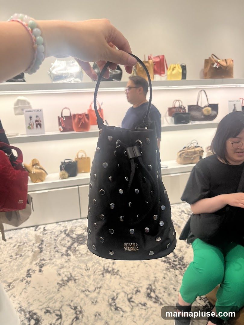 A hand holds up a black bucket bag adorned with silver studs. The bag has a drawstring closure and a braided handle. In the background, shelves display various handbags in different colors and styles. Two people are visible in the background, one man looking to the side and a woman sitting down. The store has a modern interior with white shelves and a patterned carpet. The brand name "BIMBA Y LOLA" is visible on the bag.