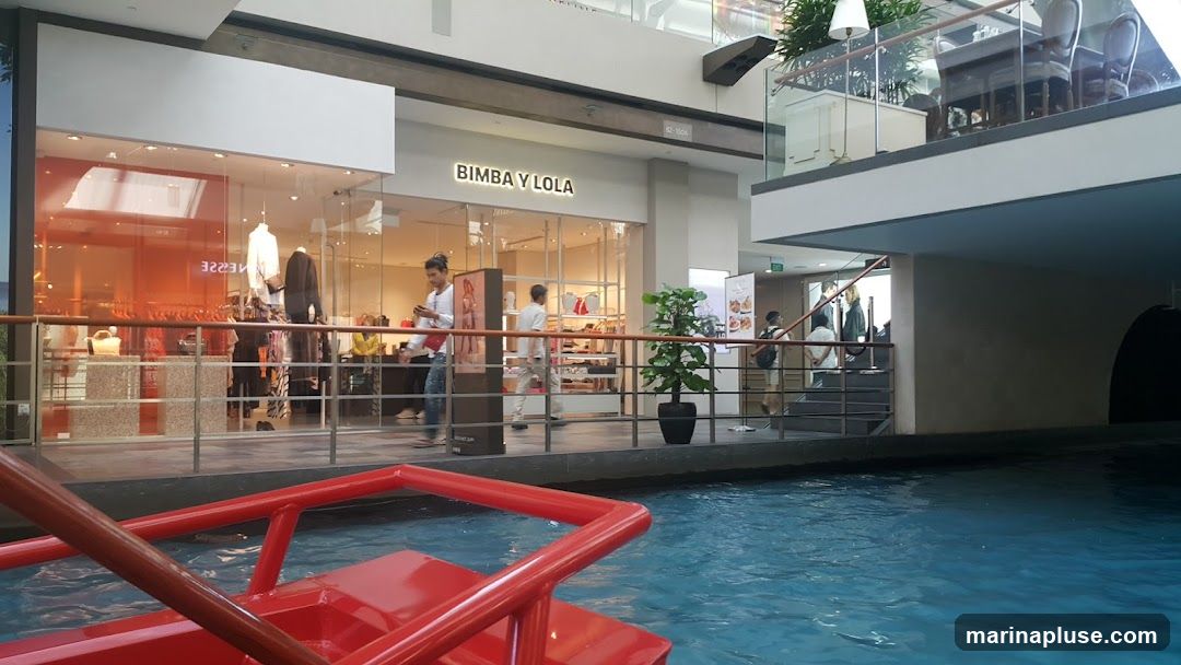 An indoor canal with blue water in the foreground, reflecting the surrounding architecture. On the right, a red railing is visible. In the background, a multi-level shopping mall interior is seen. On the upper level, a dining area with tables and chairs is set up. On the lower level, several shops are visible, including one with the prominent signage "BIMBA Y LOLA". People are seen walking around the shops and on the walkways.