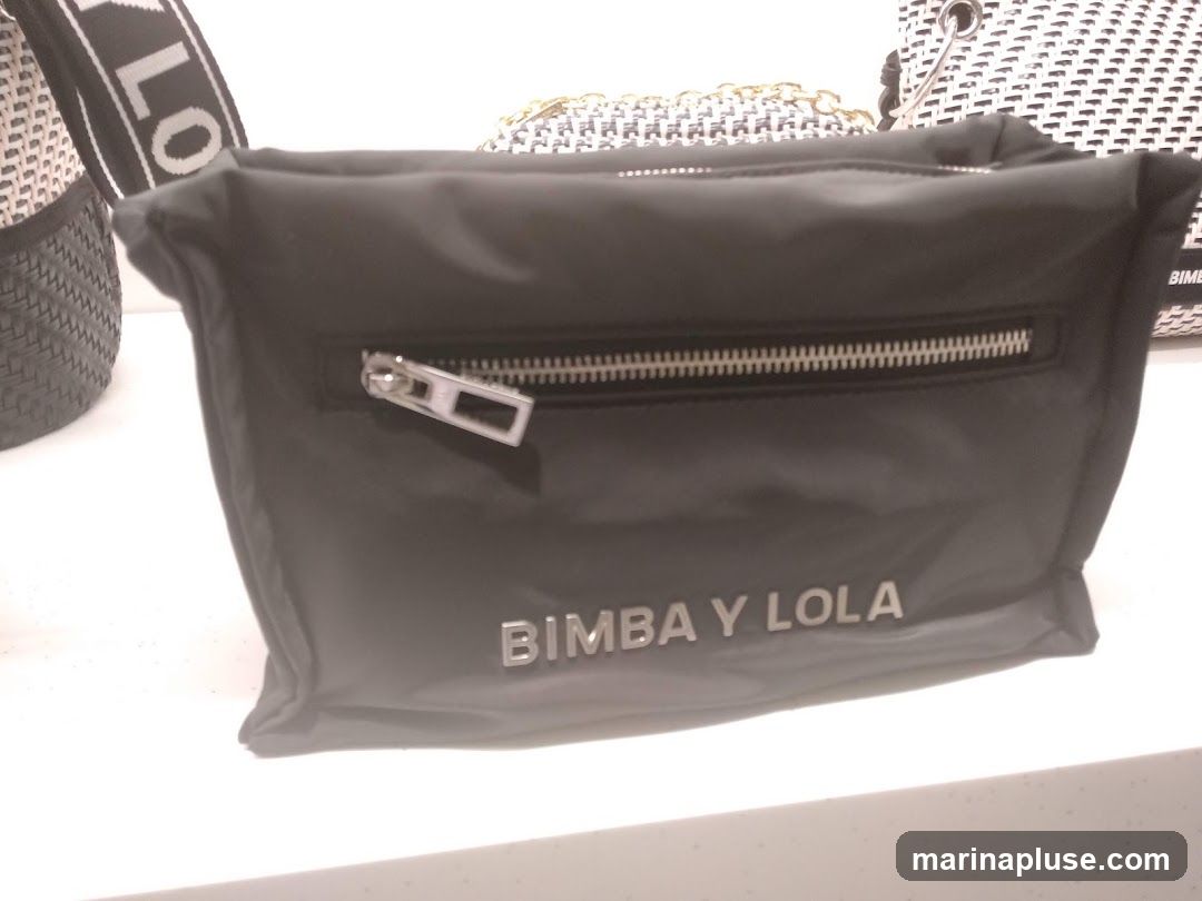 A black nylon handbag with a silver zipper is displayed on a white surface. The brand name "BIMBA Y LOLA" is embossed in silver letters on the front of the bag. Other bags, including one with a woven pattern and a strap with the word "LOVE" visible, are partially visible in the background.