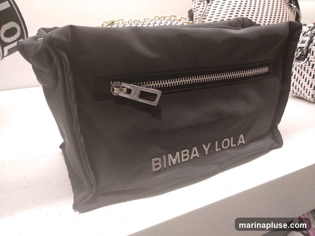 A black nylon handbag with a silver zipper is displayed on a white surface. The brand name "BIMBA Y LOLA" is visible in silver lettering on the front of the bag. Other bags, including one with a houndstooth pattern and a woven white bag, are visible in the background.