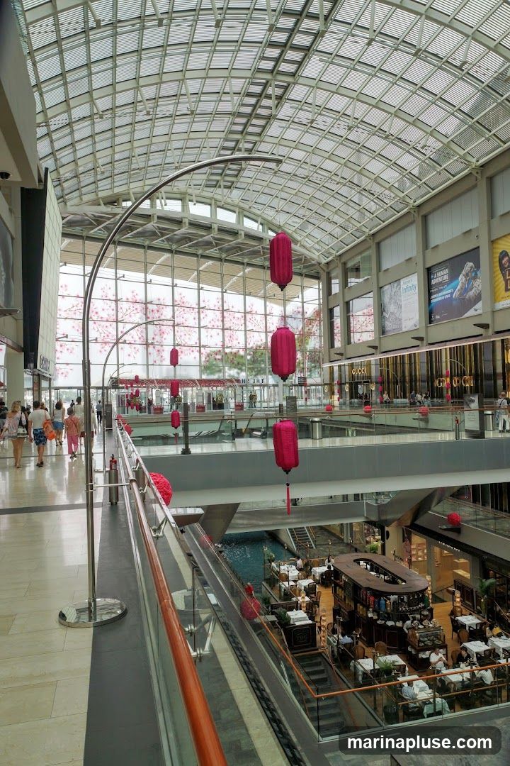 An interior view of a modern shopping mall with a glass ceiling. Red lanterns are hung as decorations. People are seen walking on the upper level, and a restaurant with tables and chairs is visible on a lower level, situated around a water feature. Storefronts with signage are visible on the right side.