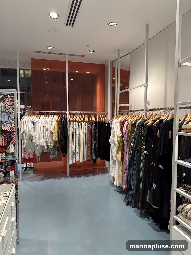 A well-lit interior of a clothing store with racks of apparel. The floor is a light blue, and the walls are white. In the background, there is an orange-tinted glass partition. Clothing items are displayed on white metal racks, featuring a variety of styles and patterns. A display case with items is visible on the left.