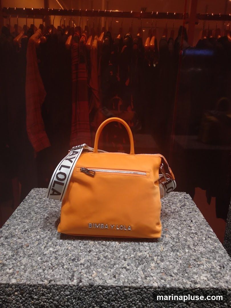 An orange handbag with a white strap featuring the text "BIMBA Y LOLA" is displayed on a speckled grey surface. Behind the handbag, clothing items hang on a rack, suggesting a retail environment. The brand name "BIMBA Y LOLA" is visible on the front of the handbag.