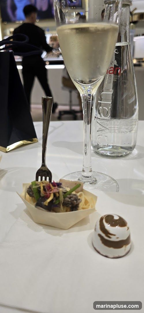 A small appetizer in a wooden boat-shaped dish with a fork inserted, a glass of champagne, and a bottle of Evian water are on a white table. A person is blurred in the background.