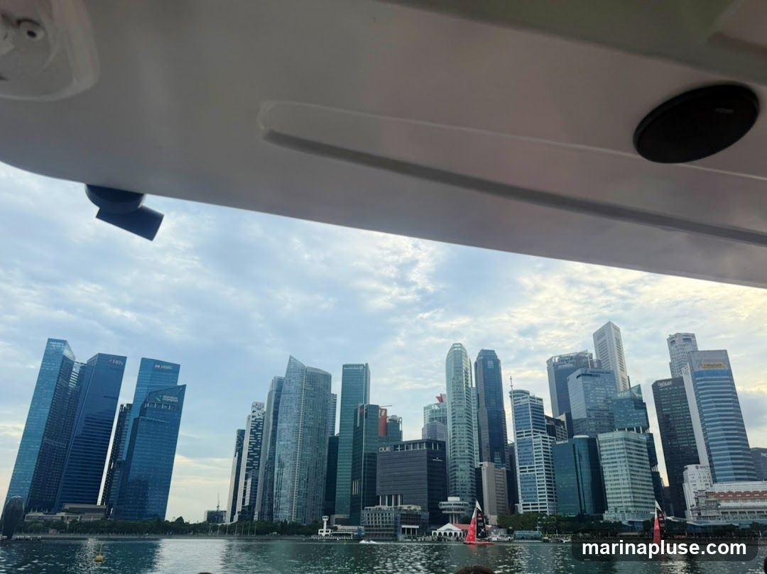 A view of the Singapore skyline with numerous modern skyscrapers, seen from the water. The buildings are a mix of glass and steel, with various architectural styles. The sky is partly cloudy. Part of a boat's interior is visible in the foreground.