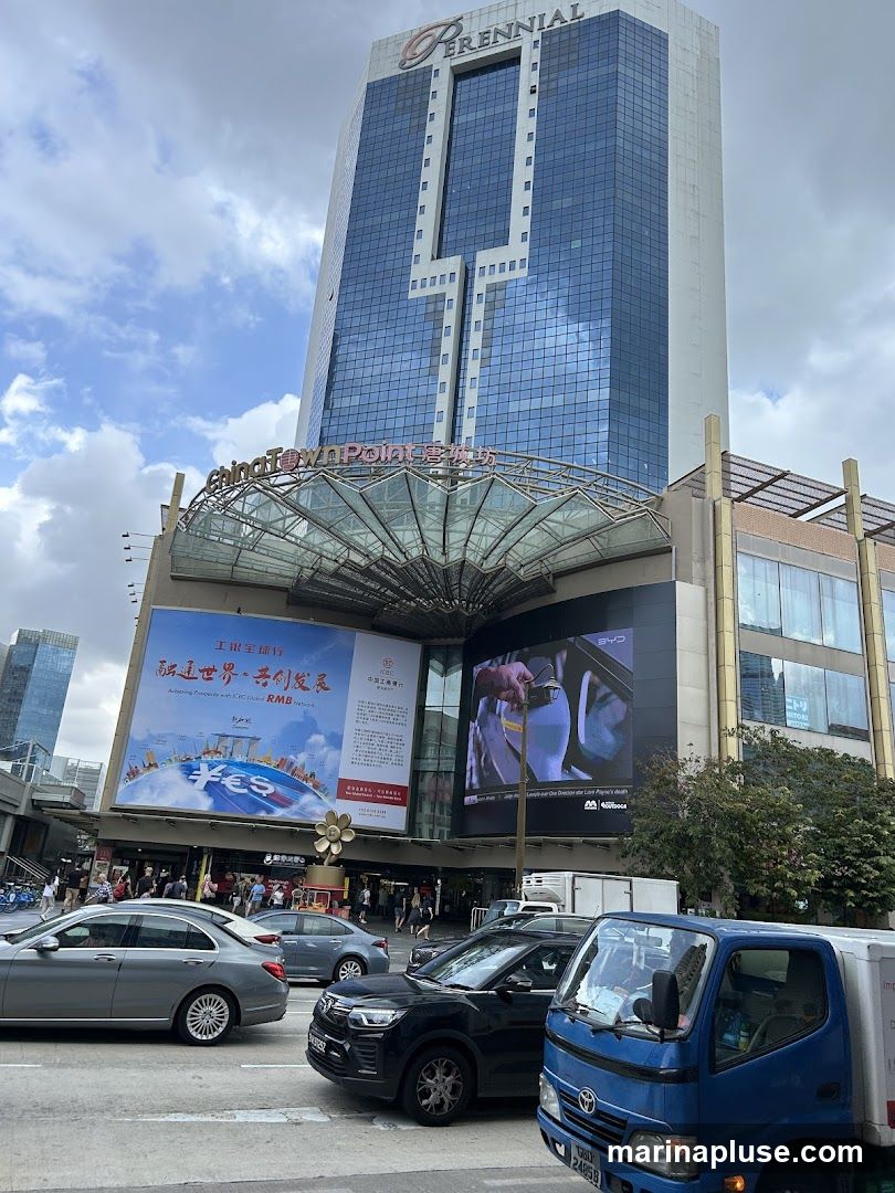 A view of the Perennial building and Chinatown Point complex from a street level. The Perennial logo is visible at the top of the skyscraper. Below it, the words "Chinatown Point" are displayed. Large billboards are mounted on the facade of the lower building, one featuring a globe with currency symbols and text in Chinese and English, and another displaying a video advertisement. Cars and a truck are on the street in the foreground.