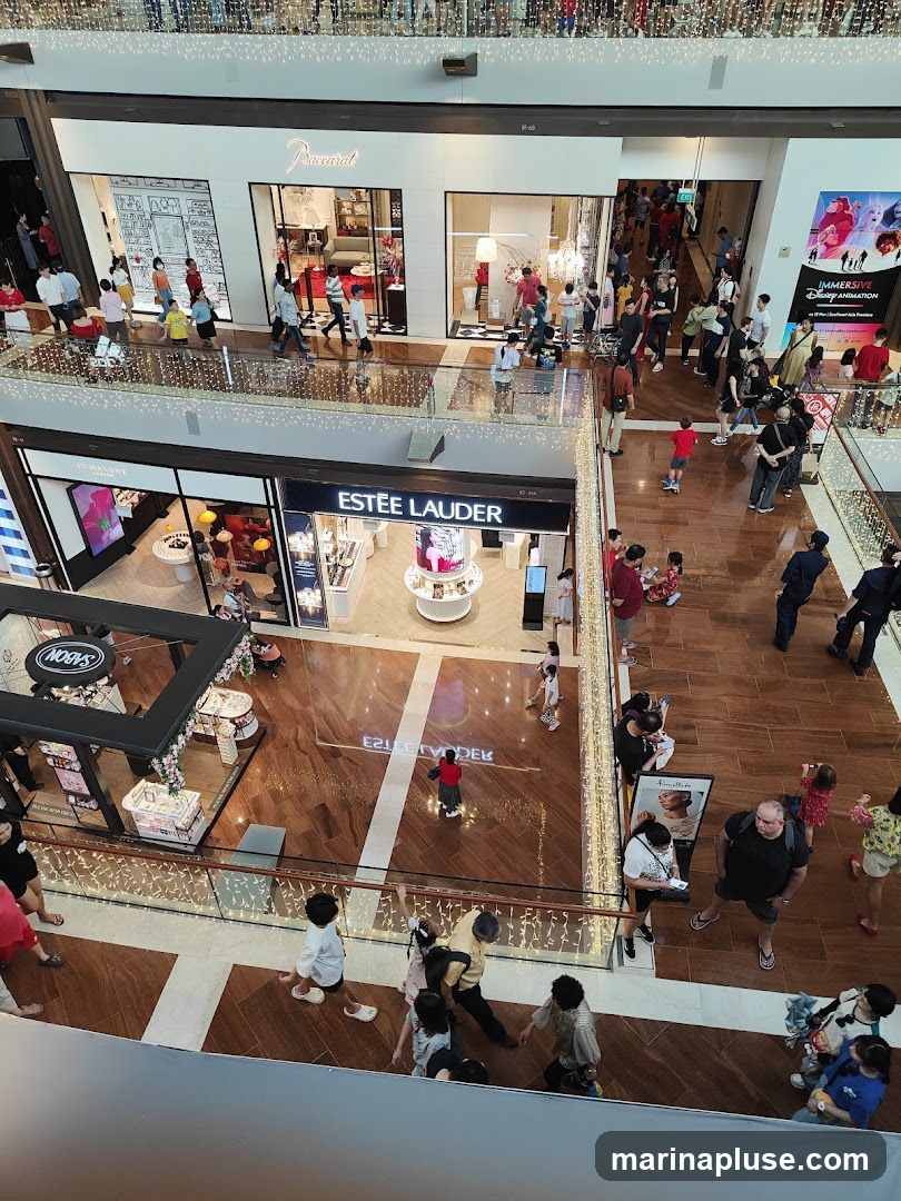 An overhead view of a busy shopping mall with multiple levels. Stores like Baccarat and Estée Lauder are visible, along with various shoppers browsing and walking around. Decorative lights adorn the railings and ceiling.