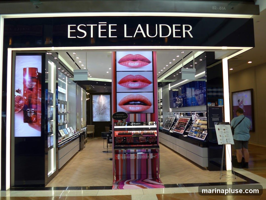 The exterior of an Estee Lauder cosmetics store. The prominent signage above the entrance reads "ESTĒE LAUDER". Inside, displays showcase various makeup products, including lipsticks and skincare. Digital screens feature images of lips and product advertisements. A person is visible browsing near a display stand.