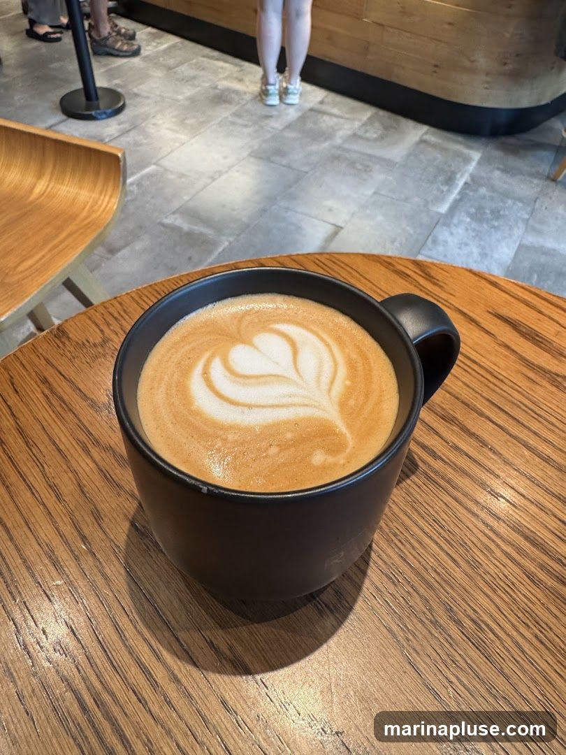 A black mug filled with a latte, featuring latte art in the shape of a heart, sits on a wooden table. In the background, people are standing in line, and a counter is visible.