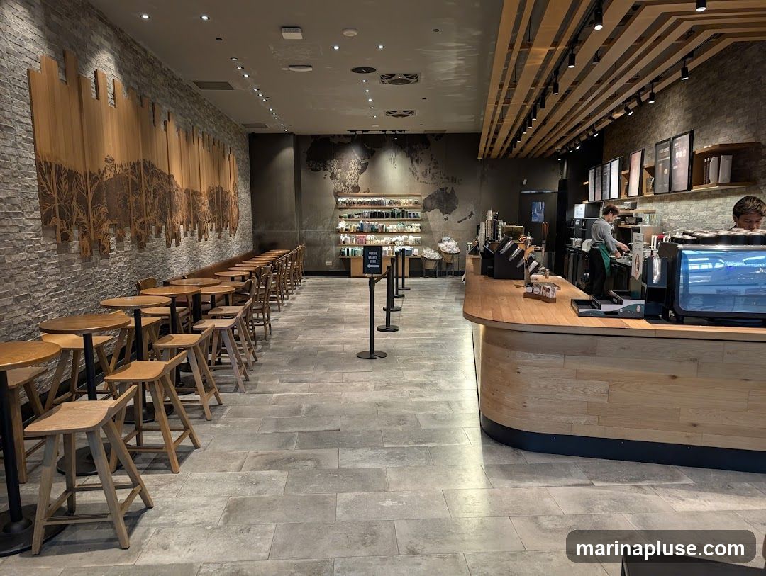 The interior of a coffee shop with seating areas and a counter. The left side of the image shows a long row of tables and stools against a brick wall decorated with a large wooden art piece. The right side features a wooden counter with baristas preparing drinks. Shelves with merchandise and a world map are visible on the back wall.
