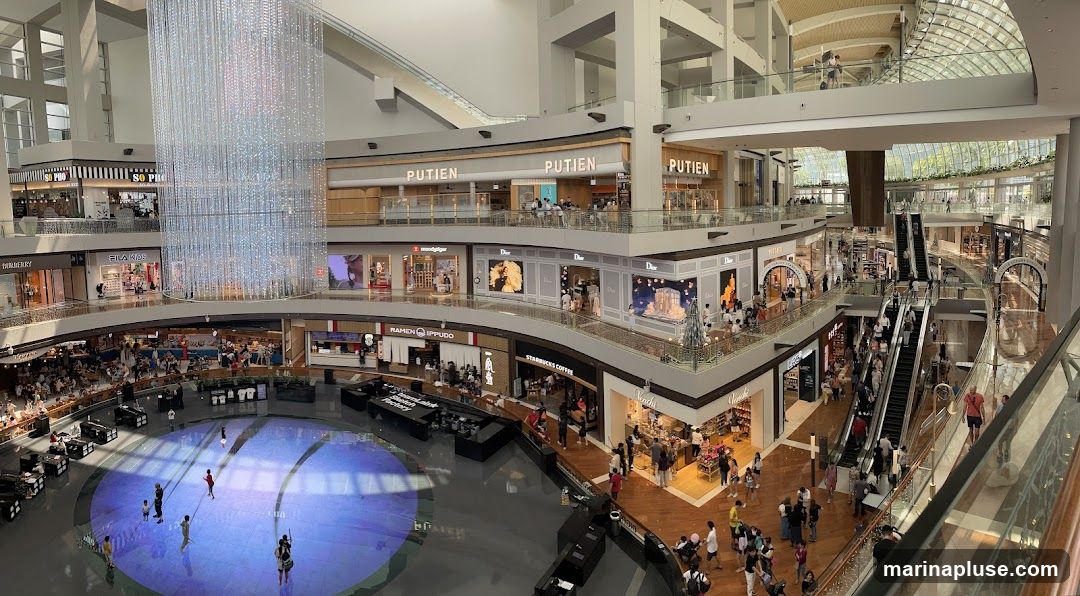 The image shows the interior of a large, modern shopping mall. A grand, multi-tiered chandelier hangs in the center of the atrium. Shops such as Dior, Starbucks Coffee, Ramen Ippudo, Fila Kids, and Mulberry are visible on various levels. Escalators and walkways connect the different floors, and many shoppers are seen browsing and moving around. The mall features a large, circular water feature or reflective surface in the center of the ground floor.