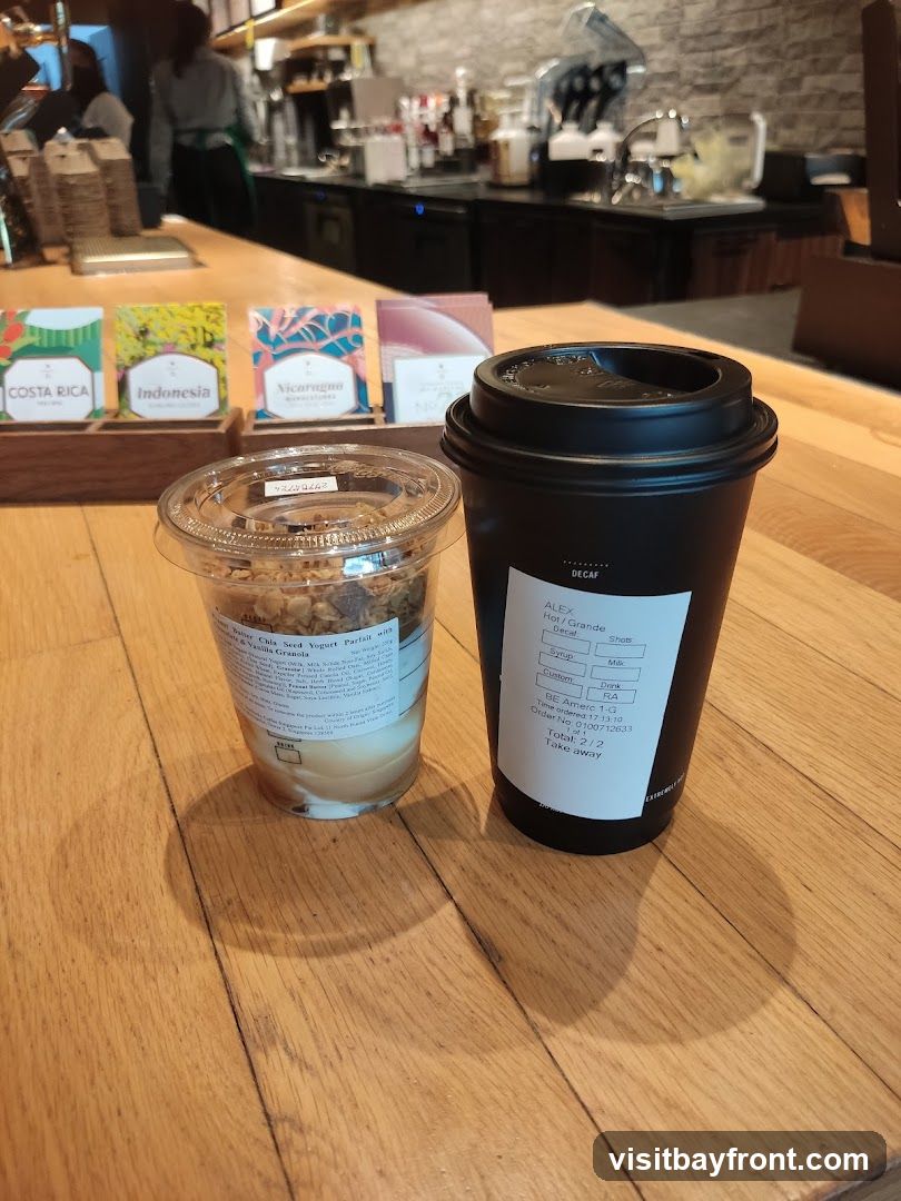 A black coffee cup and a clear cup of yogurt parfait with granola sit on a wooden counter. Behind them, a coffee shop counter with baristas and coffee machines is visible. Small colorful boxes labeled "Costa Rica," "Indonesia," and "Nicaragua" are also on the counter.