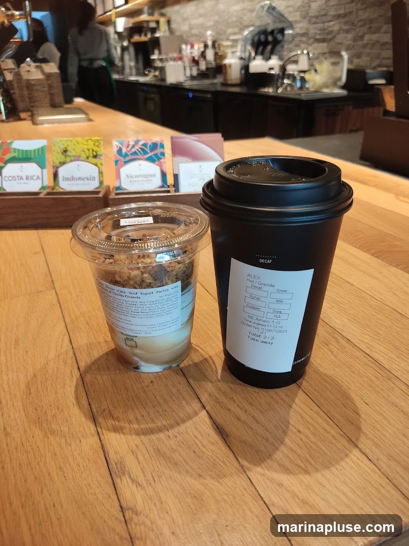 A black coffee cup and a clear cup of yogurt parfait with granola sit on a wooden counter. Behind them, a coffee shop counter with baristas and coffee machines is visible. Small colorful boxes labeled "Costa Rica," "Indonesia," and "Nicaragua" are also on the counter.