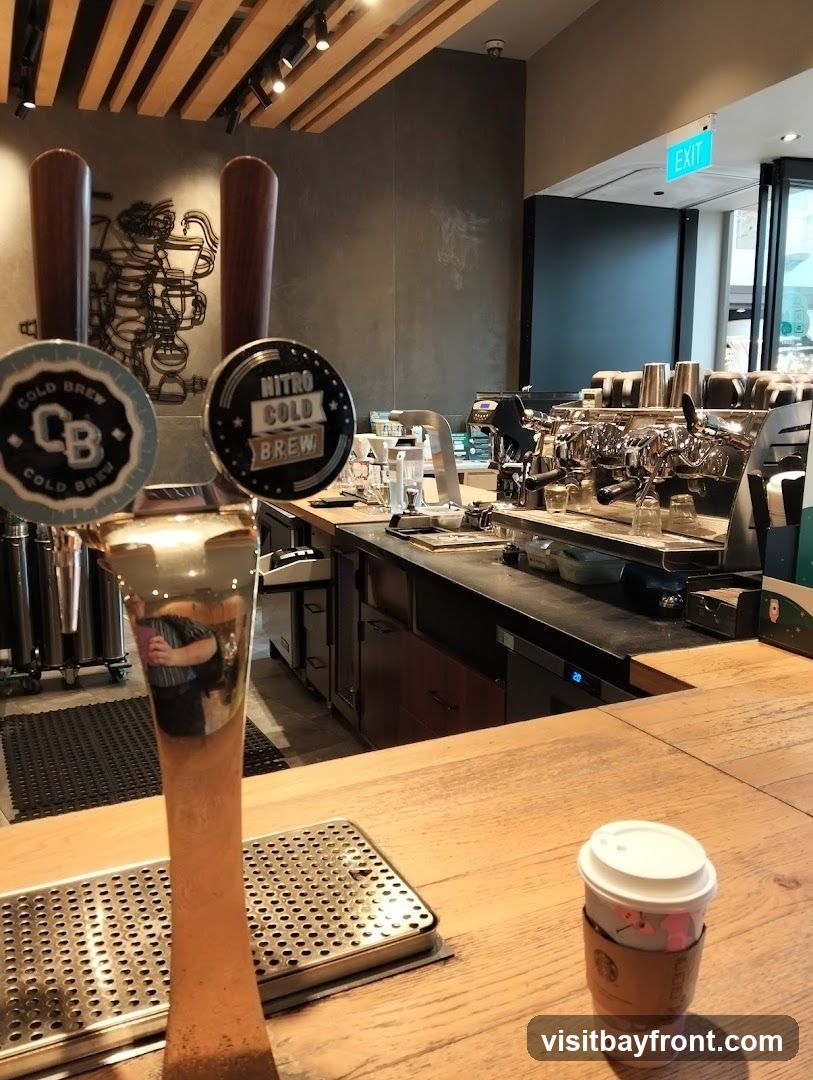 An interior view of a coffee shop, focusing on the counter area. Two taps for cold brew are prominent in the foreground, one labeled 'CB COLD BREW' and the other 'NITRO COLD BREW'. Behind the taps, a commercial espresso machine and various coffee-making equipment are visible. A cup with a lid and a branded sleeve sits on the wooden counter. An 'EXIT' sign is illuminated in green above a doorway in the background. The wall behind the counter features a decorative metal art piece depicting coffee-making apparatus.