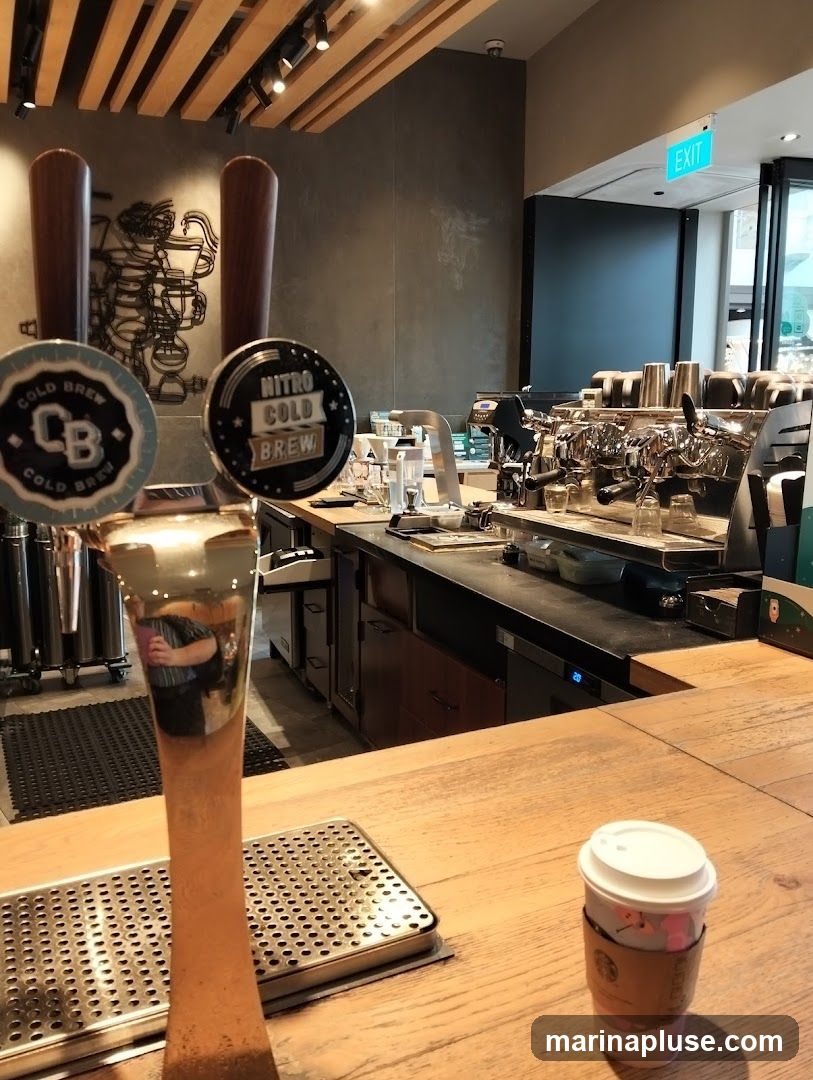 An interior view of a coffee shop, focusing on the counter area. Two taps for cold brew are prominent in the foreground, one labeled 'CB COLD BREW' and the other 'NITRO COLD BREW'. Behind the taps, a commercial espresso machine and various coffee-making equipment are visible. A cup with a lid and a branded sleeve sits on the wooden counter. An 'EXIT' sign is illuminated in green above a doorway in the background. The wall behind the counter features a decorative metal art piece depicting coffee-making apparatus.