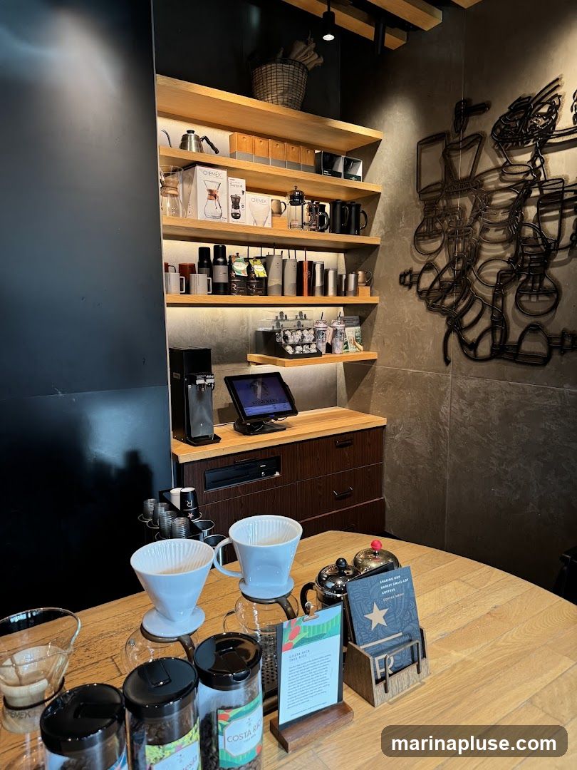 A wooden table displays coffee brewing equipment, including pour-over drippers, carafes, and canisters of coffee beans. Behind the table, shelves are stocked with various coffee-making tools, mugs, and travel tumblers. A digital display screen is visible on a counter, and decorative metal art depicting coffee-making apparatus adorns the wall.