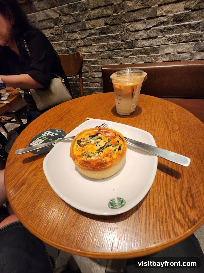 A quiche and an iced coffee are on a wooden table. The quiche is on a white plate with a Starbucks logo. The iced coffee is in a clear plastic cup with a lid. A woman is visible in the background. The wall behind the table is made of stone.