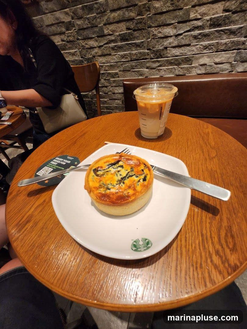 A quiche and an iced coffee are on a wooden table. The quiche is on a white plate with a Starbucks logo. The iced coffee is in a clear plastic cup with a lid. A woman is visible in the background. The wall behind the table is made of stone.