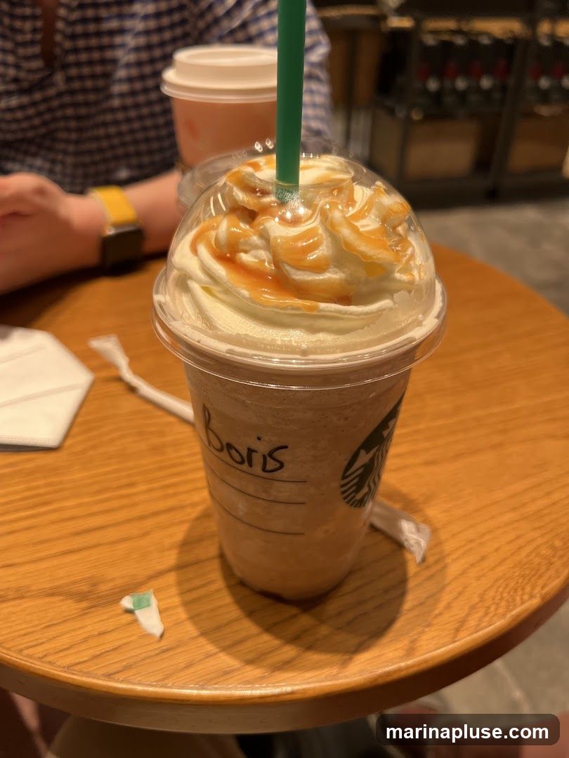 A close-up shot of a caramel frappuccino with whipped cream and caramel drizzle, with the name "Boris" written on the cup. The Starbucks logo is visible on the cup. The drink is on a wooden table, with another drink and a person's arm in the background.