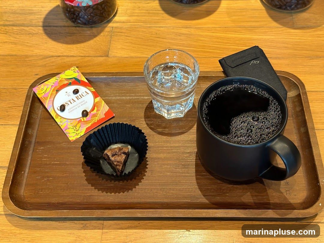A wooden tray holds a black mug of coffee, a glass of water, a small brownie in a black wrapper, and a colorful card with coffee beans. The card reads "COSTA RICA COOPVECTORIA ESENCIA DE MUJER". A small black pouch with a logo is also on the tray. In the background, jars of coffee beans are visible.