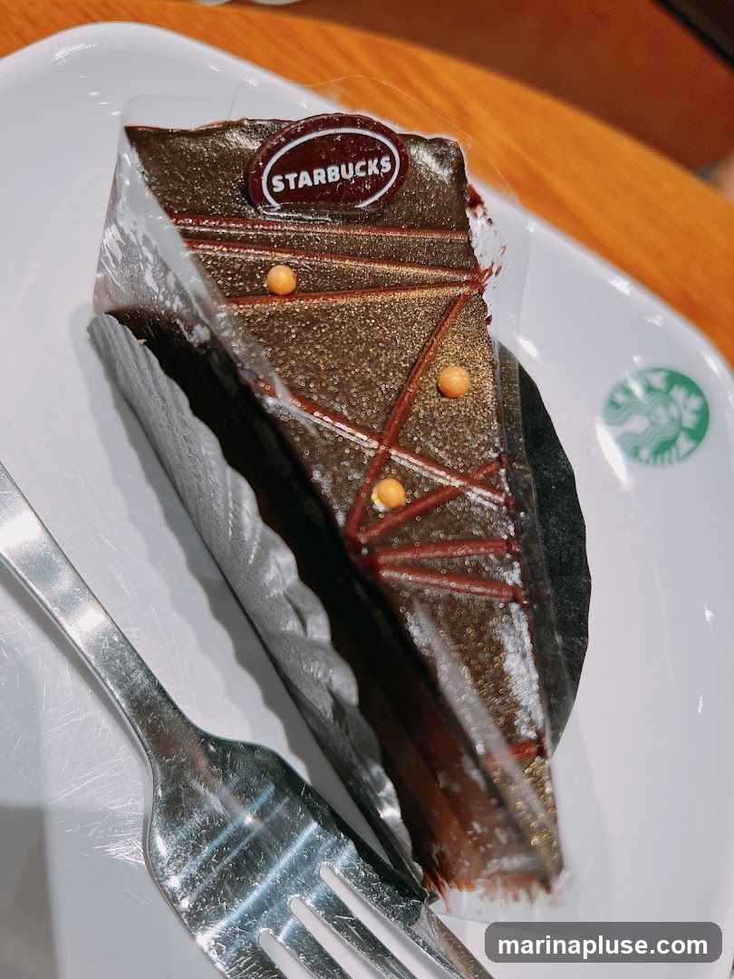 A slice of chocolate cake with a Starbucks logo on top, served on a white plate with a fork. The cake is decorated with chocolate icing, red lines, and small orange balls. The plate also has a faint Starbucks logo.