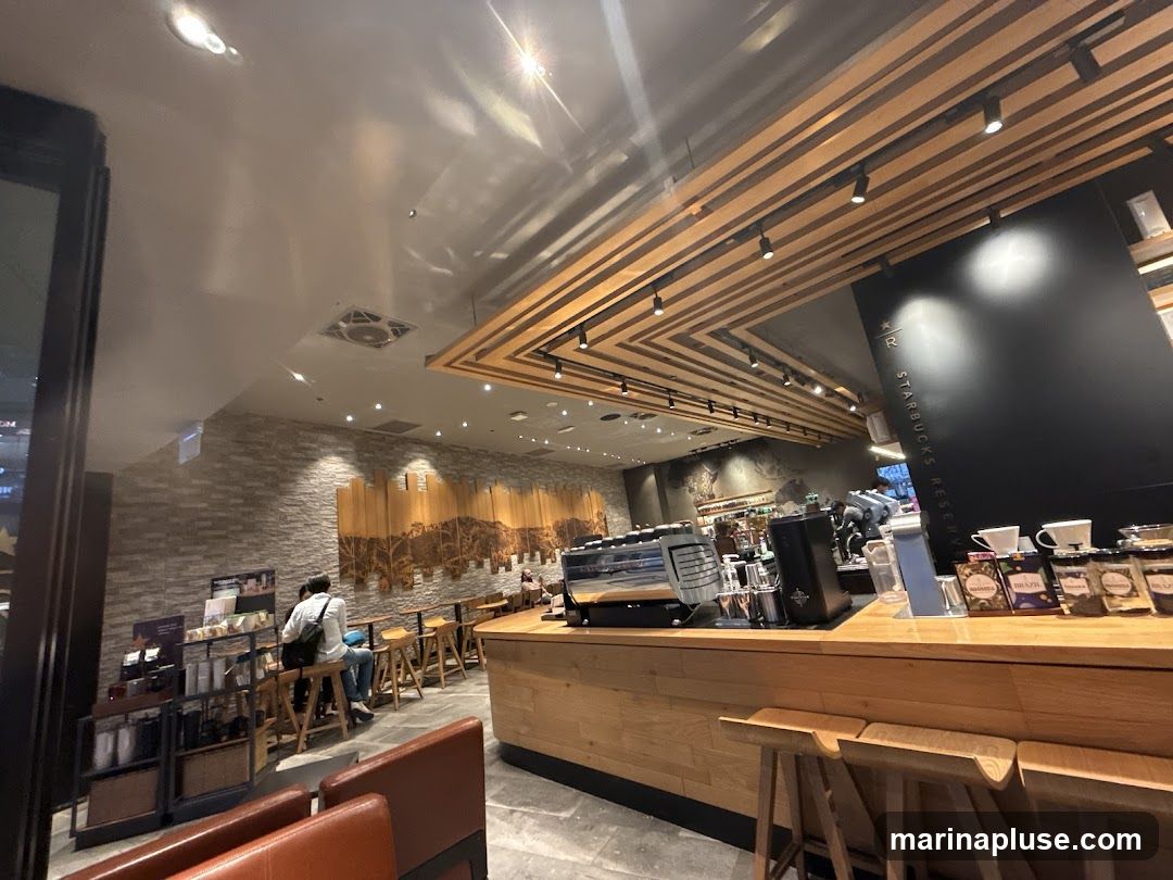 The interior of a Starbucks Reserve cafe with a modern design. The ceiling features wooden slats and track lighting. The walls are decorated with a textured stone pattern and a large wooden mural. A counter area displays coffee machines, cups, and packaged coffee beans. Several patrons are seated at high-top tables and a bar.