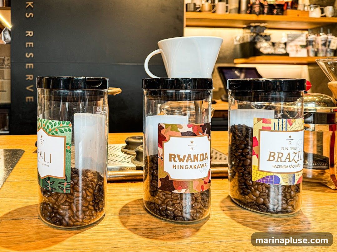 Three clear jars filled with coffee beans are displayed on a wooden counter. Each jar has a label indicating the origin of the beans: Bali, Rwanda Hingakawa, and Sun-Dried Brazil Fazenda São João. Behind the jars, a black sign with "KS RESERVE" is visible, along with shelves stocked with mugs and coffee-making equipment.