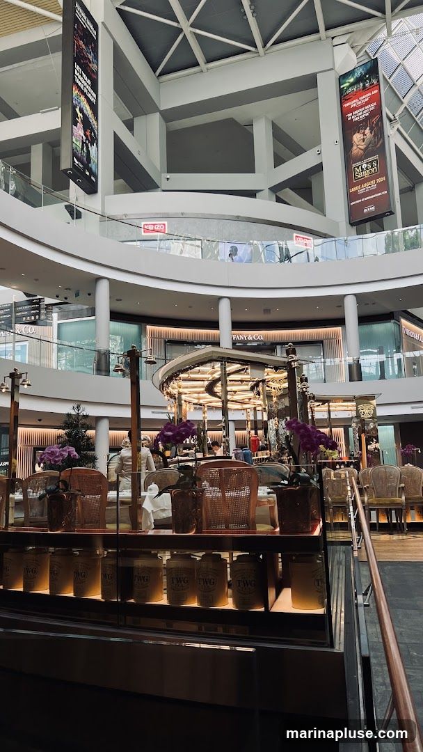The image shows the interior of a modern shopping mall with multiple levels. In the foreground, there is a cafe with tables and chairs, and a display case filled with jars labeled "TWG TEA". In the background, various shops are visible, including Tiffany & Co. and Kenzo. Large digital billboards hang from the ceiling, displaying advertisements.
