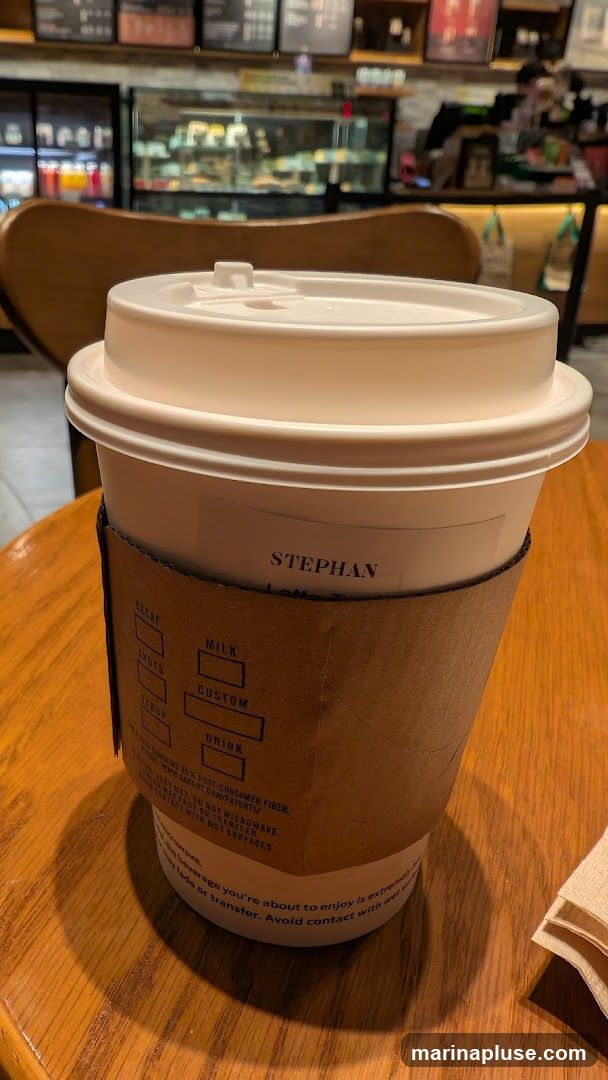 A close-up of a white coffee cup with a brown cardboard sleeve. The name