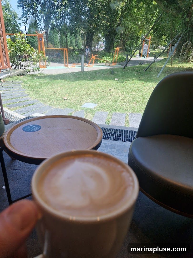 A close-up of a latte with heart art in a white mug, held by a hand. In the background, through a window, is an outdoor seating area with a round wooden table and a dark leather chair. Beyond that, a grassy area with trees and outdoor exercise equipment is visible.