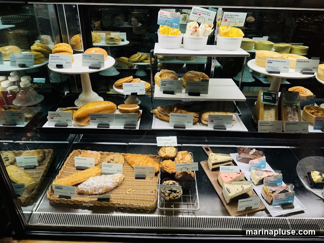 A refrigerated display case filled with various baked goods and pastries. Items include bagels, scones, croissants, muffins, cookies, cakes, and pies. Some items have price labels.