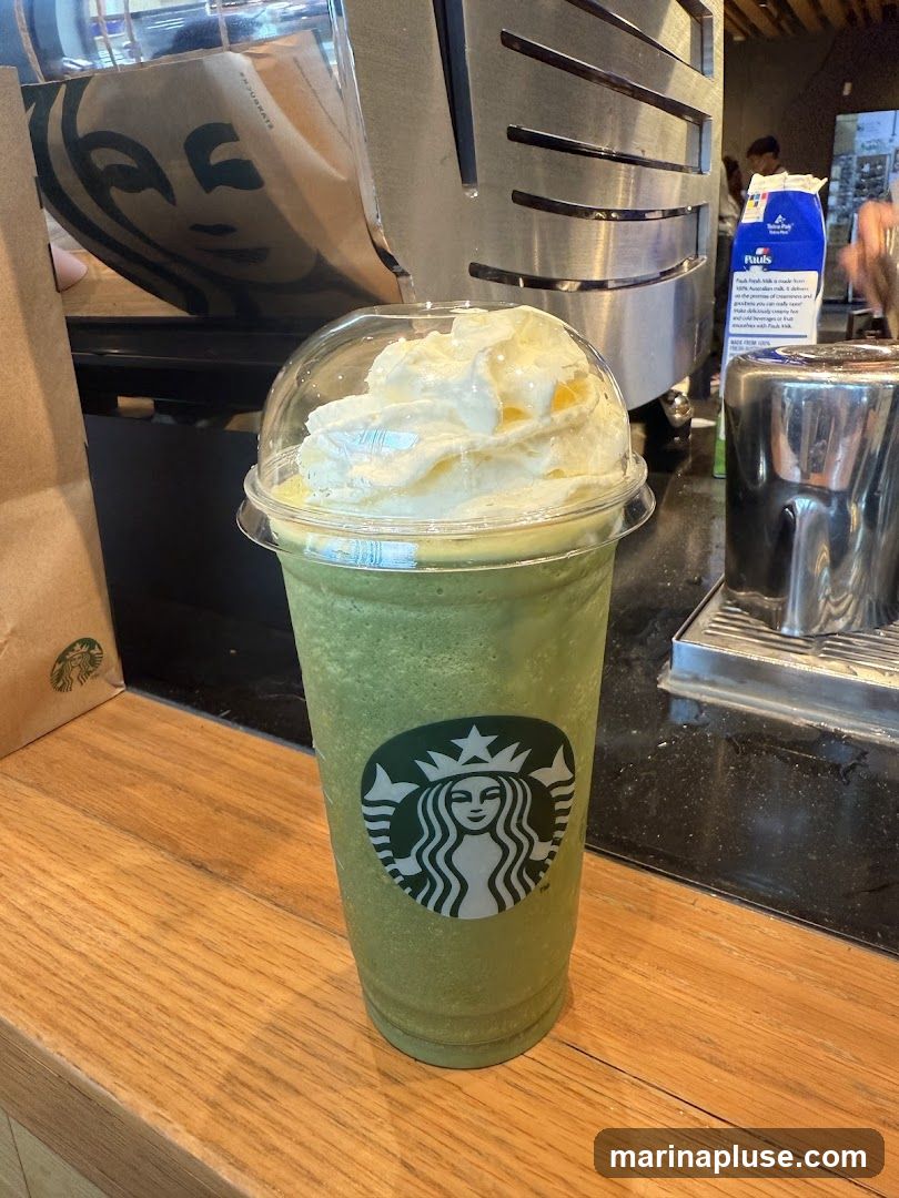 A green blended beverage, likely a matcha Frappuccino, topped with a generous swirl of whipped cream and a sprinkle of topping. The drink is in a clear plastic cup with a domed lid and features the Starbucks logo. It sits on a wooden counter. In the background, a Starbucks paper bag with the logo is visible, along with coffee-making equipment and a carton of milk.