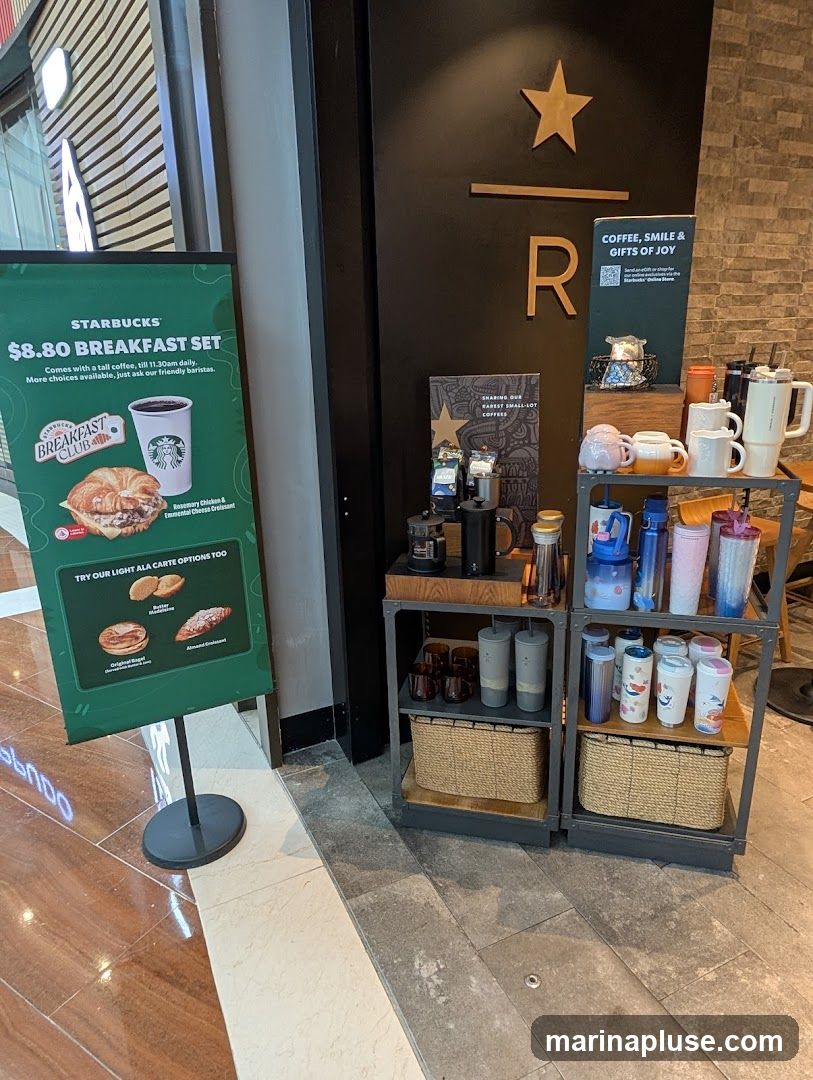 A Starbucks promotional sign displays an $8.80 breakfast set, featuring a croissant and coffee. Adjacent to the sign is a display shelf with various Starbucks merchandise, including mugs, tumblers, and coffee brewing equipment. The background shows a dark door with the Starbucks logo and a large 'R'.