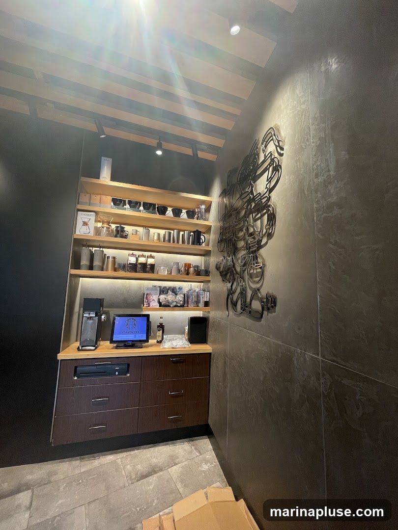 A view of a coffee shop counter with a computer monitor displaying the Starbucks logo, a coffee machine, and drawers. Above the counter are shelves stocked with coffee cups, brewing equipment, and other merchandise. A large metal art piece is mounted on the wall to the right.