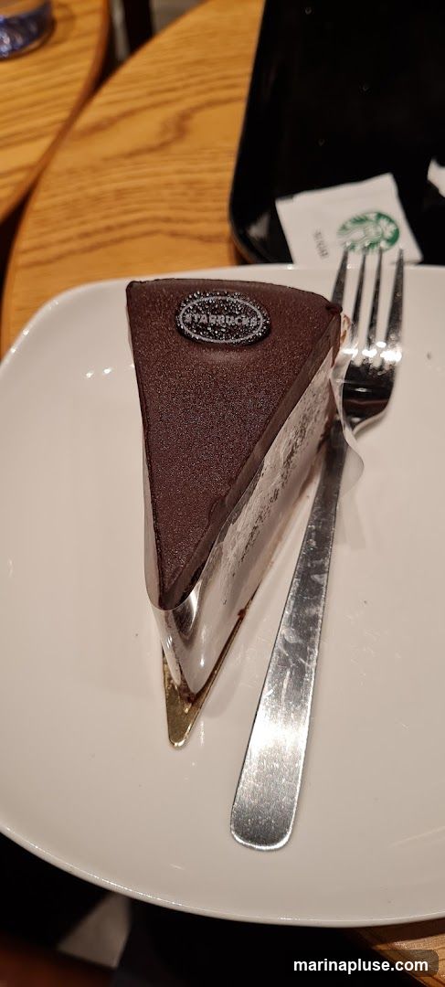A slice of chocolate cake with a dark chocolate glaze is served on a white plate with a fork and a sugar packet. The cake has a Starbucks logo on top.