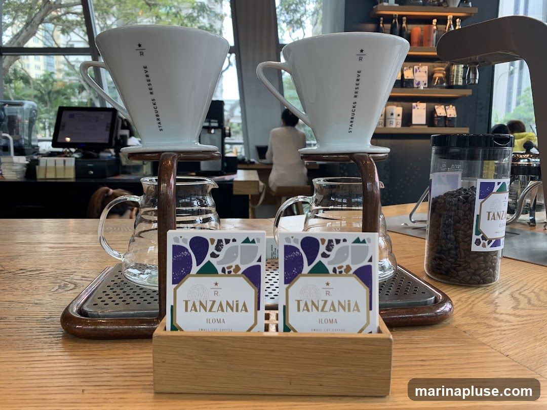 A close-up view of a coffee brewing station at Starbucks Reserve, featuring two pour-over drippers with glass carafes below. Two small cards labeled "TANZANIA ILOMA SMALL-LOT COFFEE" are placed in a wooden holder in the foreground. A clear container filled with coffee beans is visible to the right, also labeled "TANZANIA ILOMA". In the background, a barista is visible working behind the counter, with shelves displaying coffee-related items.