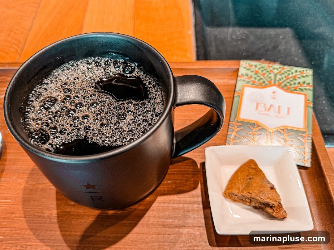 A black mug filled with dark coffee, showing a layer of bubbles on top, sits on a wooden tray. Next to the mug is a small white dish containing a triangular piece of a cookie or brownie. Behind the dish is a small, decorative packet labeled "BALI" with a green and orange pattern. The mug has a subtle logo visible on its side.