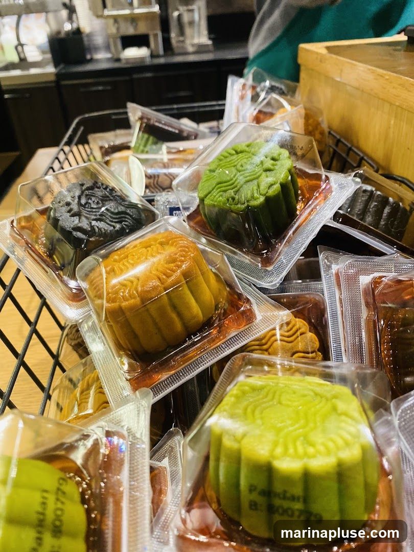 A close-up shot of several individually packaged mooncakes in a basket. The mooncakes are in various colors, including green, black, and golden brown, and feature intricate embossed patterns on top. Some packaging shows the text "Pandari B: 800779". In the background, a coffee shop counter with equipment is visible.