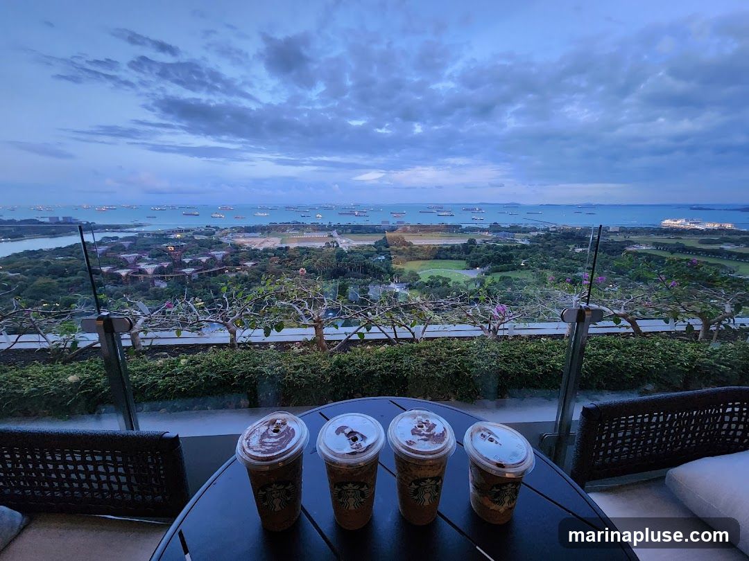 Four Starbucks drinks are on a round table overlooking a bay filled with ships. The sky is cloudy and the light suggests dusk. In the distance, there are green fields and futuristic-looking structures.