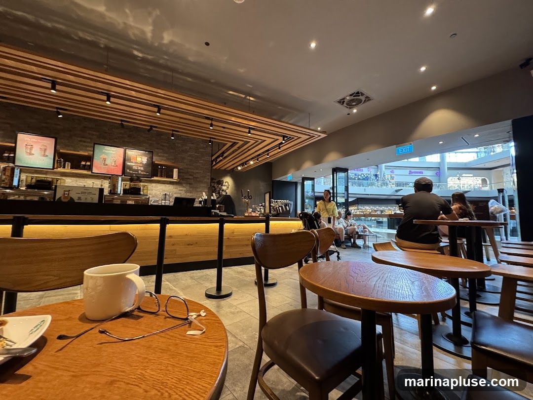 An interior view of a coffee shop with a counter, seating areas, and customers. Digital menu boards are visible above the counter, displaying drink promotions. The overall ambiance is modern and casual, with wooden accents and warm lighting. Through an open area, a shopping mall interior is visible.