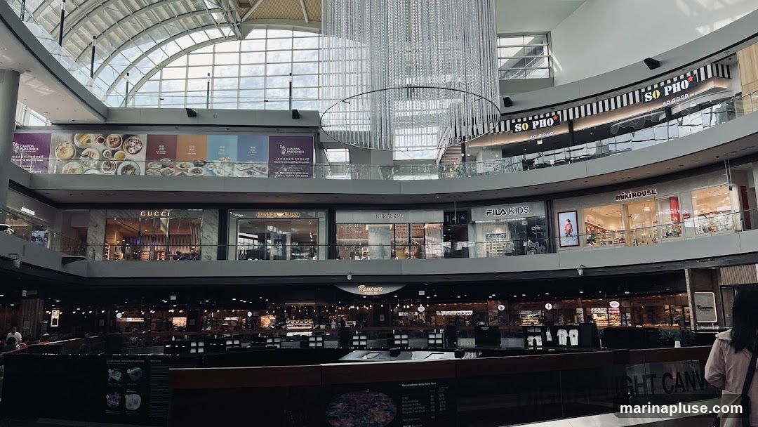 An interior view of a multi-level shopping mall. Visible stores include Gucci, Burberry, Fila Kids, Miki House, and So Pho. A large, ornate chandelier hangs from the ceiling. Banners displaying food and store promotions are visible on upper levels. The lower level appears to be a food court or dining area.