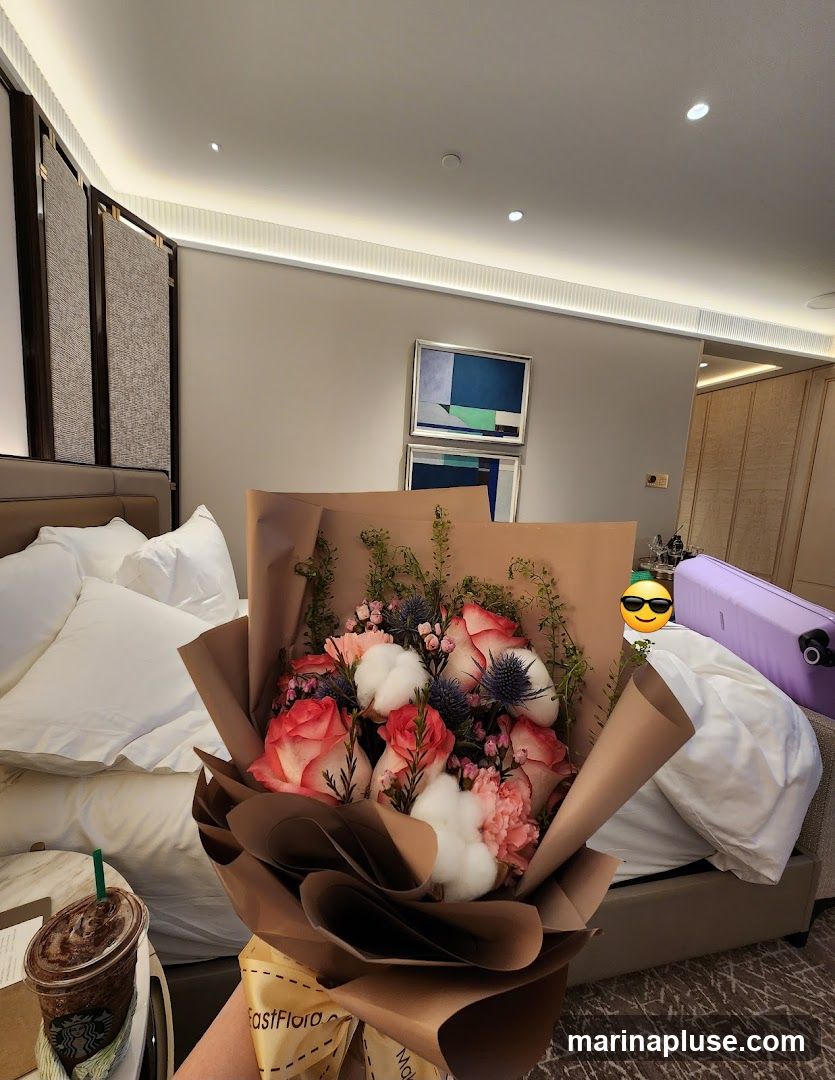 A bouquet of pink and white flowers is held in the foreground, with a Starbucks iced coffee on a side table to the left. The background shows a hotel room with a large bed, abstract art on the wall, and a purple suitcase.